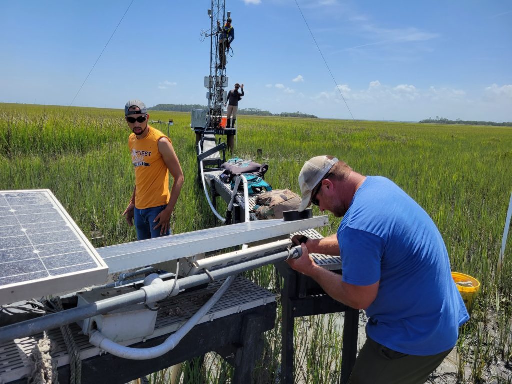 Two people working on a solar panel array on a boardwalk in the marsh. Farther along the boardwalk, two people are on and next to the flux tower.