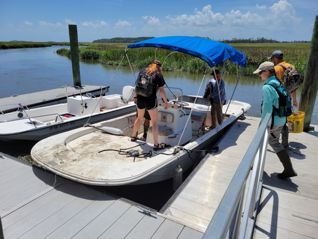 Four people loading equipment onto a small boat at a dock with marsh and tidal creek in the background